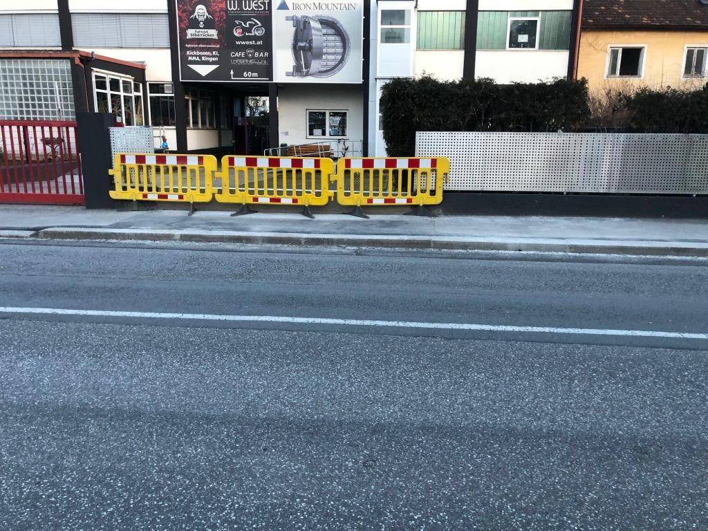 Nahaufnahme der Straßenseite eines Gebäudes mit gelben Absperrungen und Geschäftsfassaden im Vordergrund.