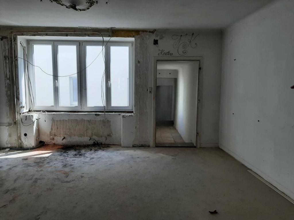 Innenraum im Sanierungszustand mit freiliegenden Kabeln, beschädigten Wänden und großen Fenstern.