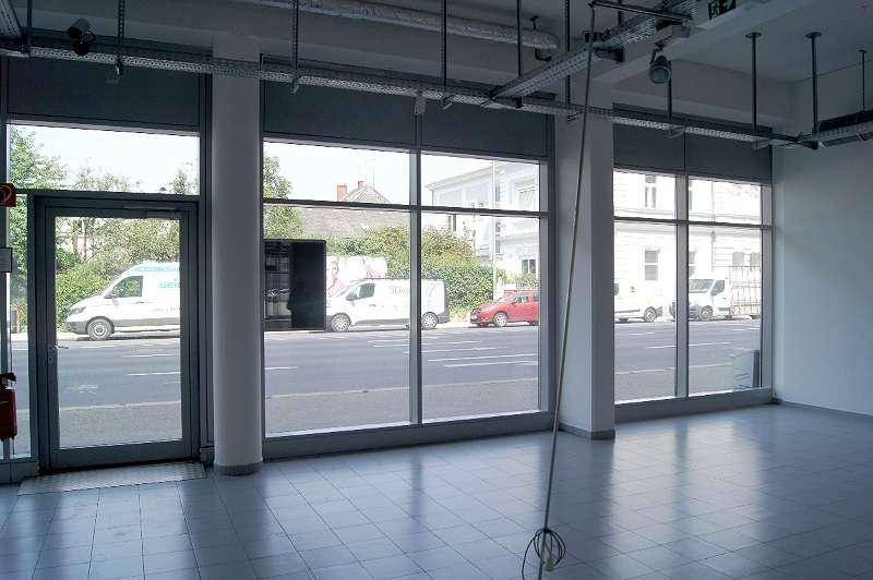 Heller Gewerberaum mit großen Schaufenstern zur Straße, Fliesenboden und sichtbaren Deckeninstallationen.