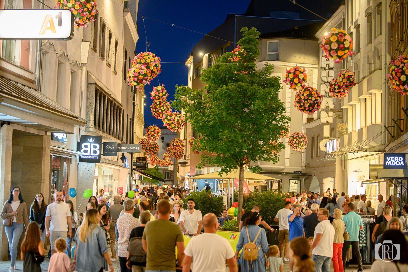 Abendliche Szene einer belebten Einkaufsstraße mit vielen Menschen, hell erleuchteten Geschäften und festlicher Dekoration.