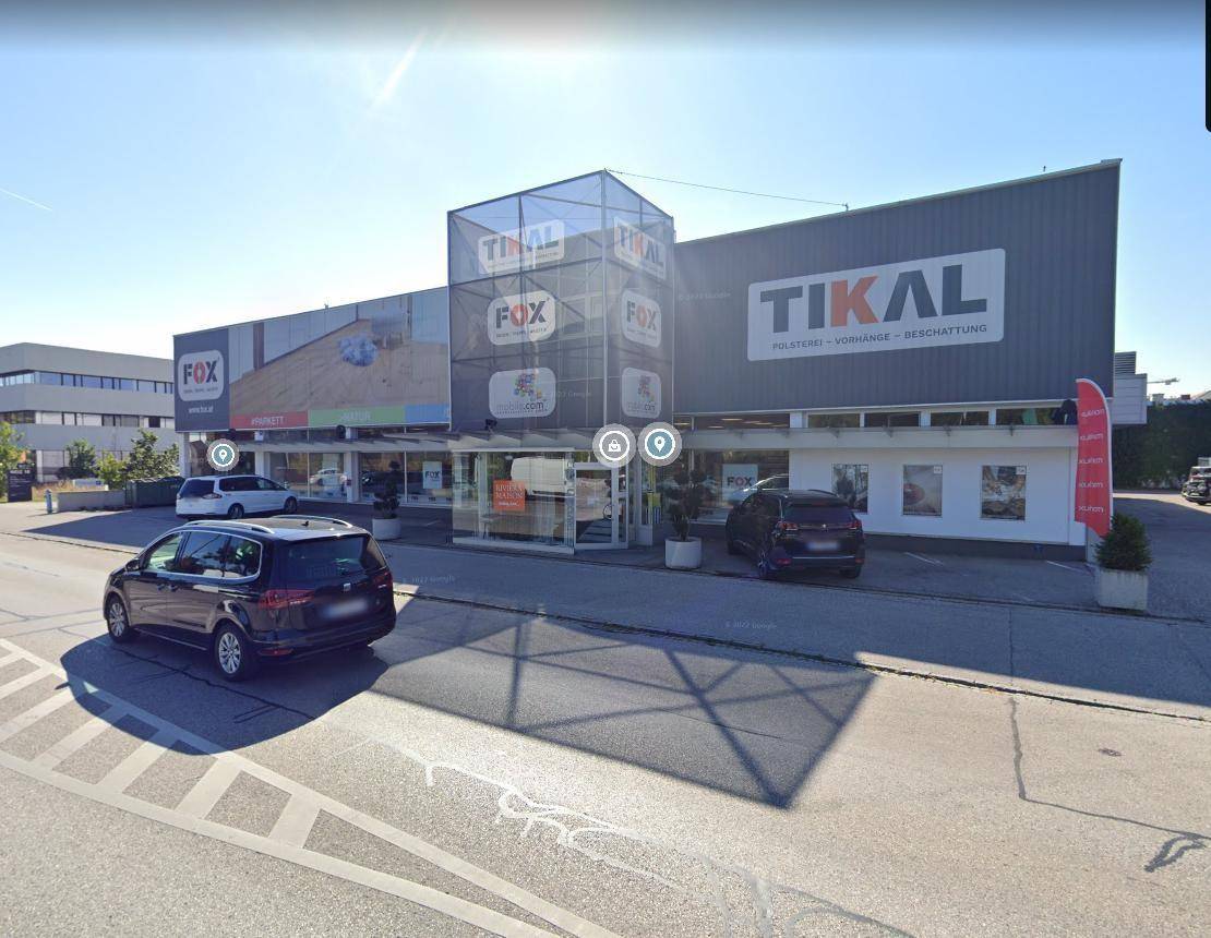 Ansicht eines Geschäftsgebäudes mit markanter Glasfassade am Eingang und umliegenden Parkflächen.
