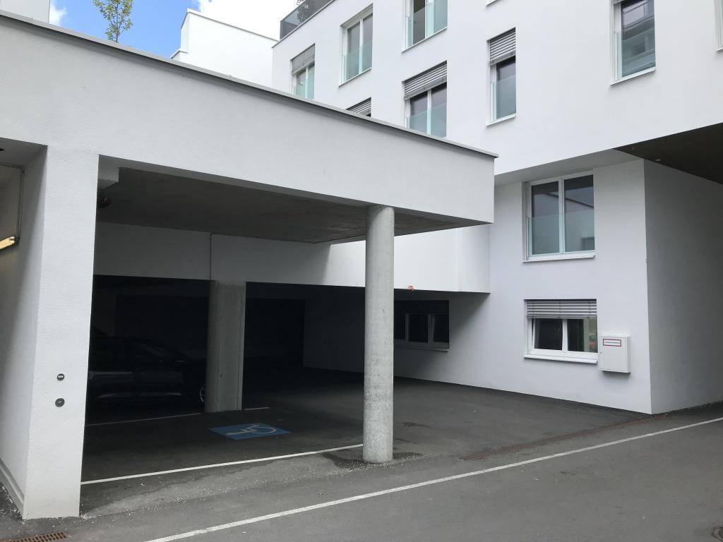 Überdachter Parkplatzbereich mit einem Behindertenparkplatz neben einem modernen weißen Gebäude.