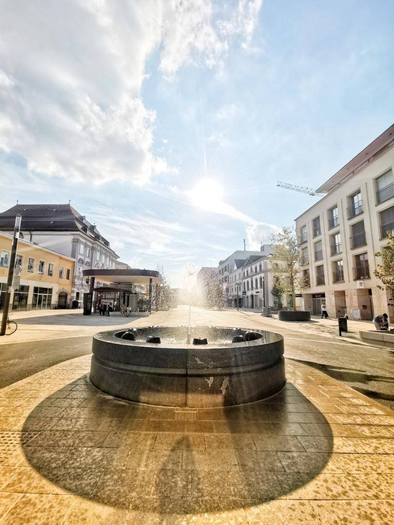 Sonnige Stadtstraße mit einem modernen Brunnen im Vordergrund und Gebäuden unter einem blauen Himmel.
