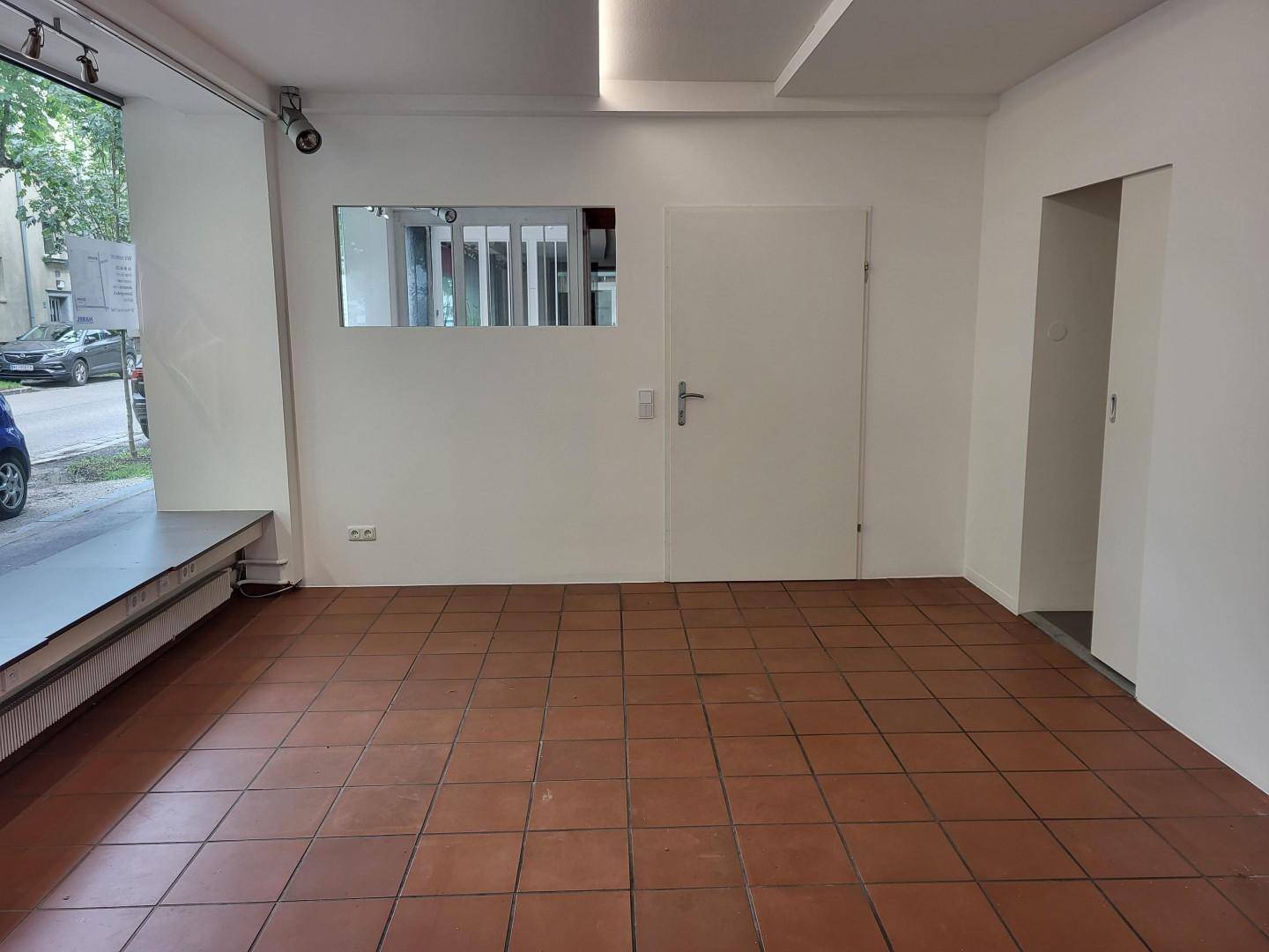 Leerer Verkaufsraum mit Terrakotta-Fliesenboden, weißen Wänden und einem kleinen Fenster zur Rückseite.