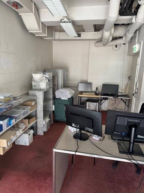 Bürofläche mit mehreren Arbeitsplätzen, Regalen und sichtbaren Lüftungsrohren an der Decke.