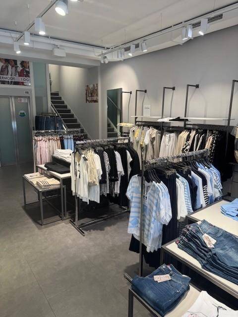 Verkaufsraum mit Treppe zum Obergeschoss, präsentiert eine Auswahl an Kleidung und Accessoires auf Fliesenboden.
