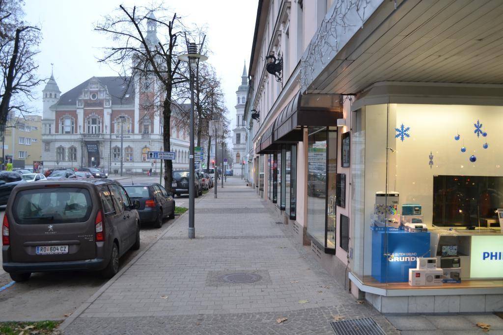 Blick auf eine belebte Straße mit parkenden Autos und Geschäftsfassaden in einer Stadt.
