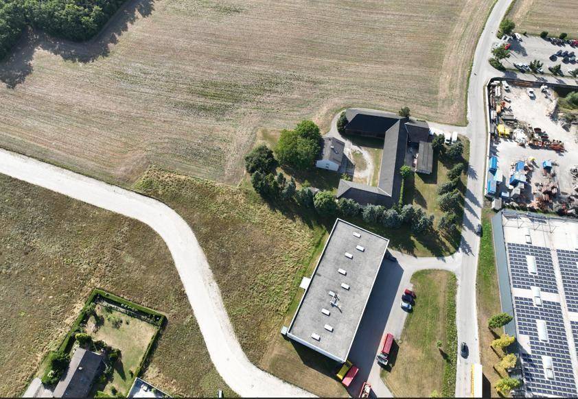 Luftaufnahme eines Gewerbegebiets mit mehreren Gebäuden, einer Zufahrtsstraße und weiten Feldern im Hintergrund.