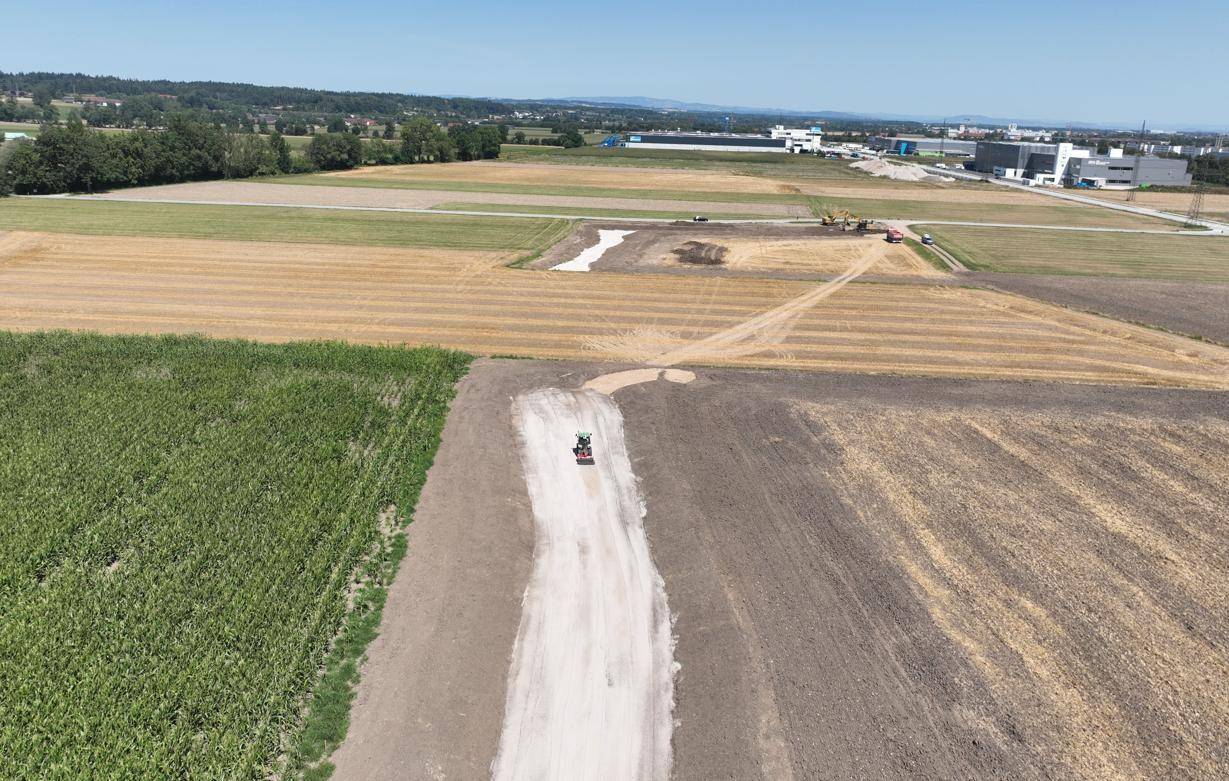 Luftaufnahme eines Baugeländes mit vorbereiteten Flächen und landwirtschaftlichen Feldern unter klarem Himmel.
