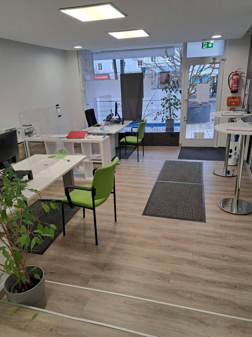 Moderner Büroraum mit großen Fenstern, hellem Laminatboden und grünen Akzentstühlen, gut beleuchtet.