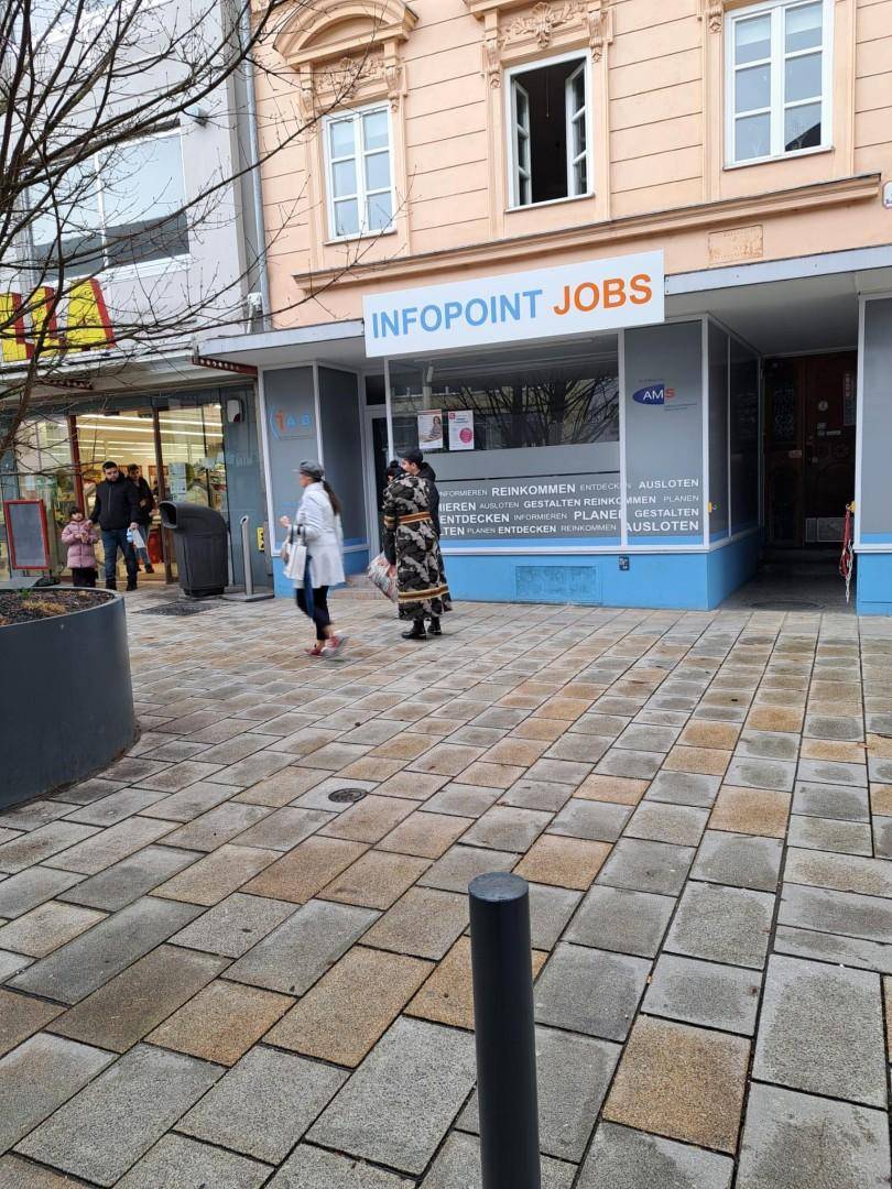 Geschäftsfassade mit Infopoint Jobs Schild und Passanten auf dem gepflasterten Gehweg.