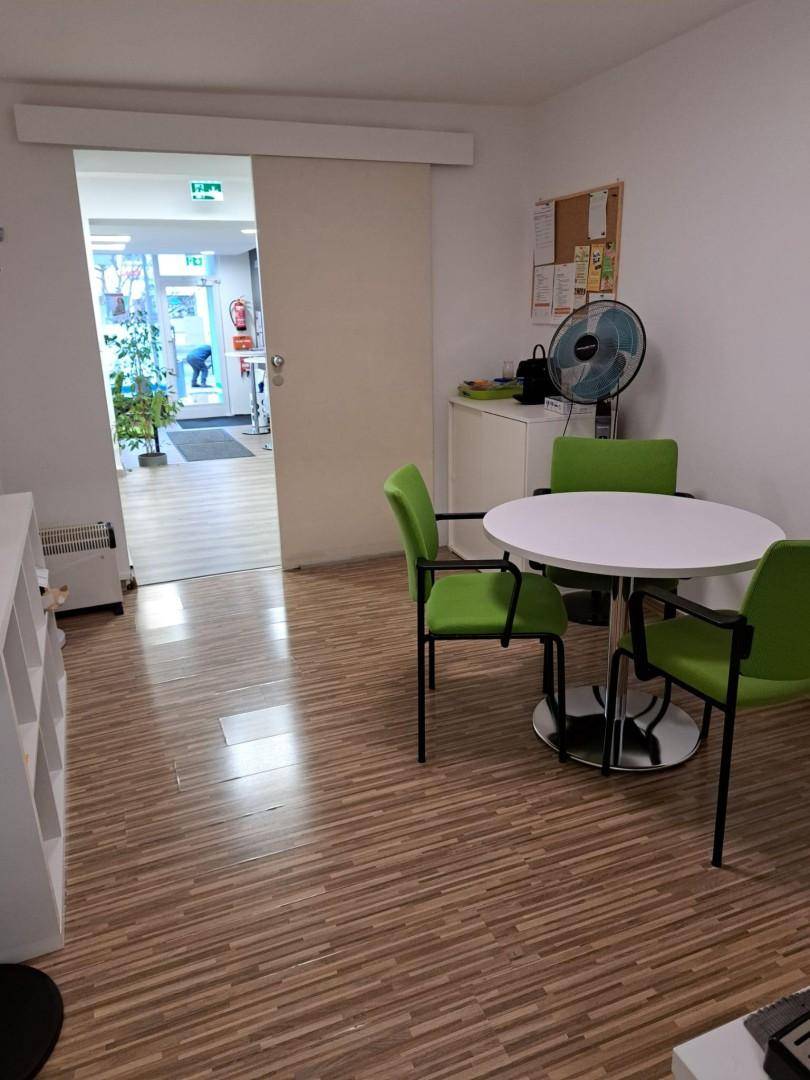 Heller Bürobereich mit kleinem Besprechungstisch und grünen Stühlen, durch Schiebetür abgetrennt.