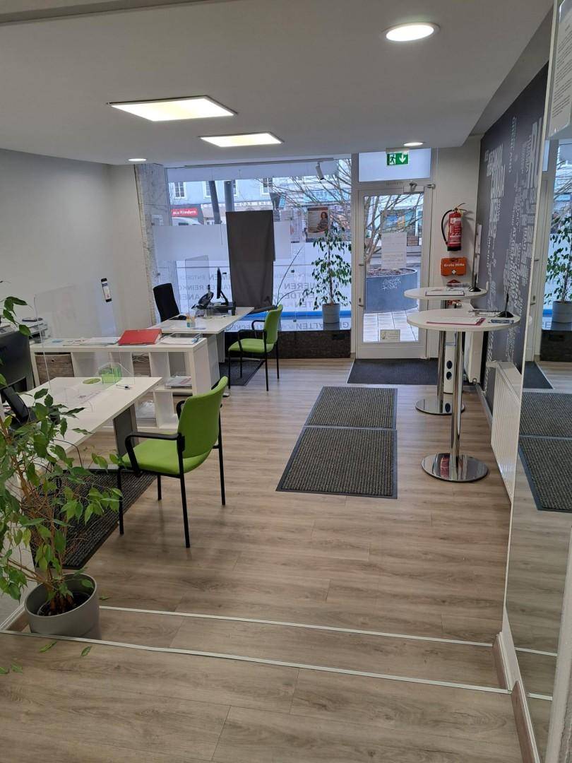 Helles Büro mit mehreren Arbeitsplätzen, grünen Stühlen und großer Fensterfront zur Straße.