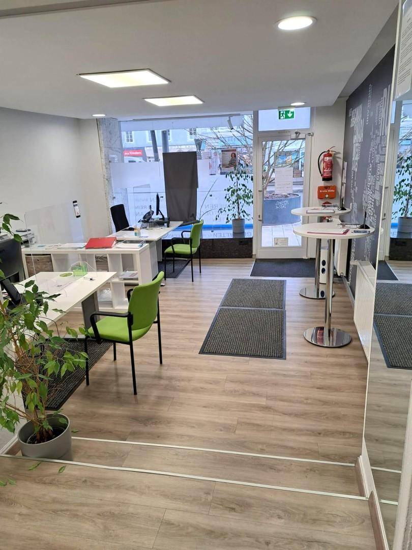 Heller Büroraum mit großen Schaufenstern, modernen Schreibtischen und grünen Bürostühlen, funktional eingerichtet.