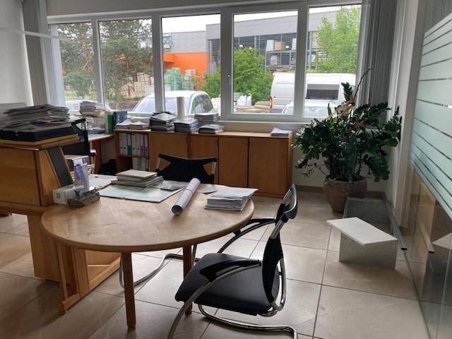 Büroraum mit Holztisch, Bürostuhl und großen Fenstern, die einen Blick auf das Außengelände bieten.