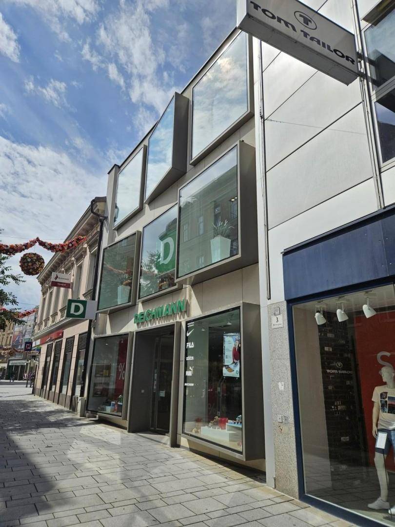 Geschäftsgebäude mit moderner Glasfassade und dem Tom Tailor Geschäft an einer sonnigen Einkaufsstraße.