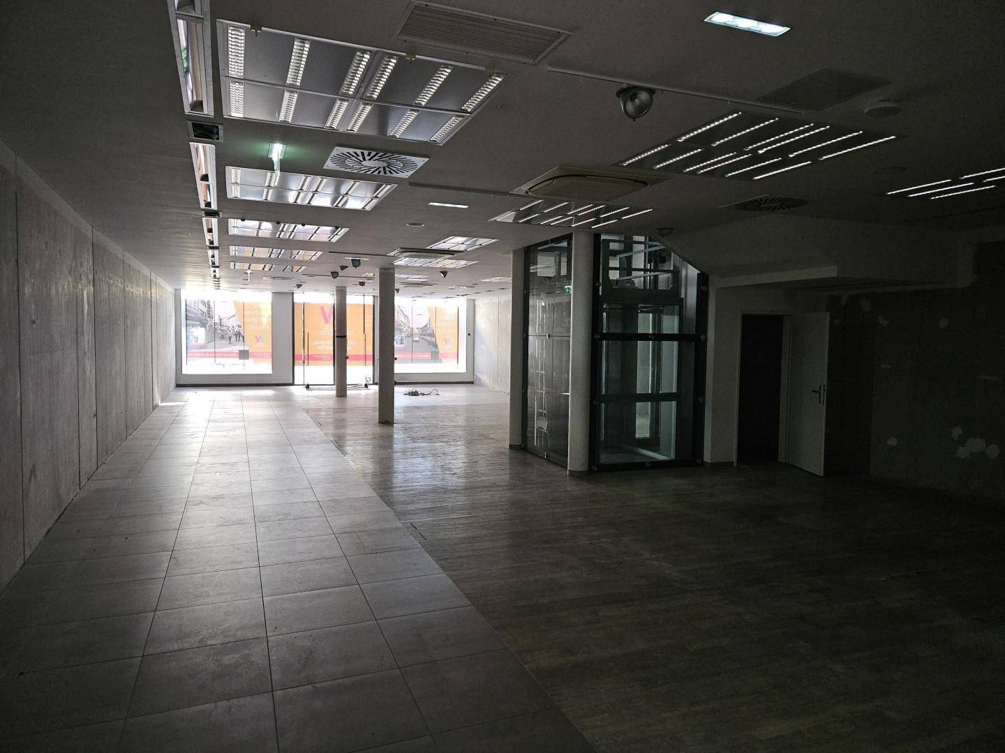 Geräumiger Gewerberaum mit Fliesenboden, Glasaufzug und Treppenaufgang im Hintergrund.