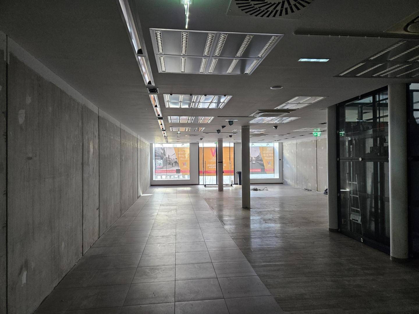 Langer, leerer Gewerberaum mit Fliesenboden, Deckenbeleuchtung und großen Schaufenstern zur Straße.