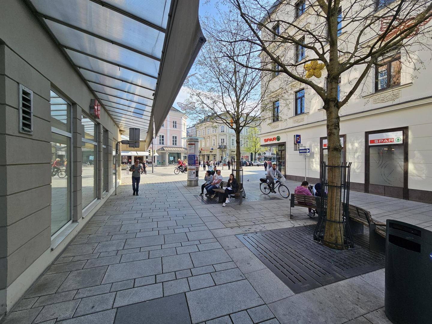 Panoramablick auf eine belebte Fußgängerzone mit Geschäften, Passanten und Bäumen unter klarem Himmel.