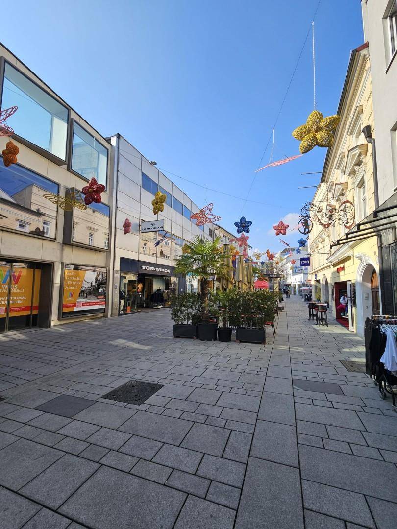 Belebtes Stadtbild einer Fußgängerzone mit Geschäften, Außengastronomie und bunten Dekorationen in der Luft.