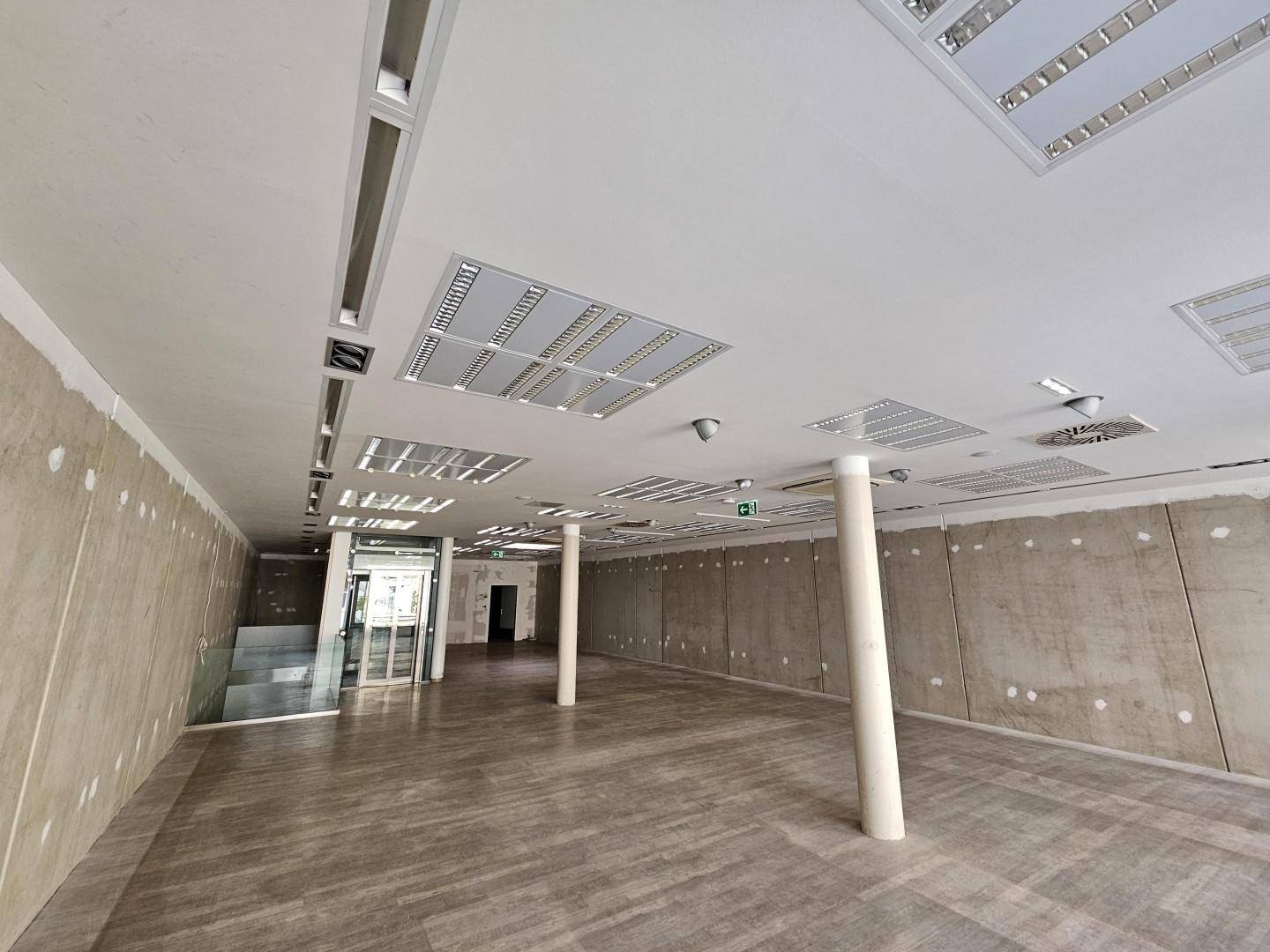Weitläufiger Gewerberaum mit Holzoptik-Boden, hohen Decken, Säulen und einem modernen Aufzugsbereich.