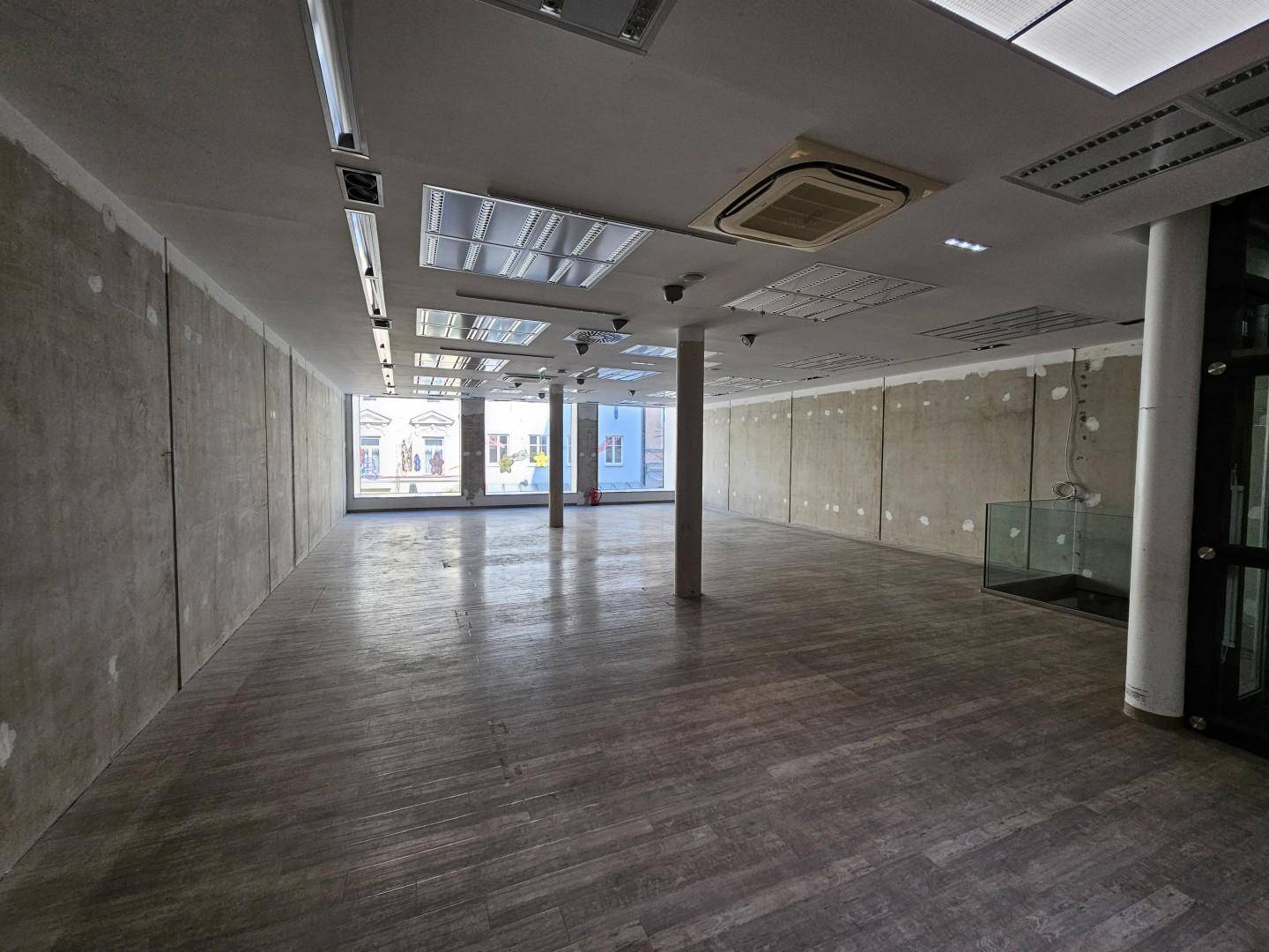 Großer, leerer Gewerberaum mit Holzoptik-Boden, hohen Decken und großen Fenstern zur Straße hin.