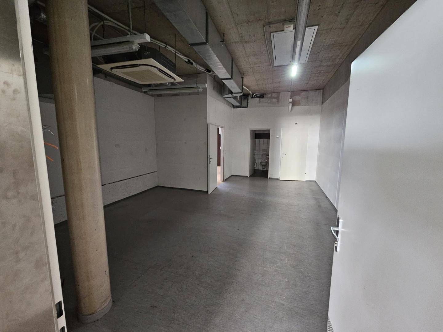 Leerer Gewerberaum mit Betonboden, sichtbaren Lüftungsrohren an der Decke und mehreren Türen.