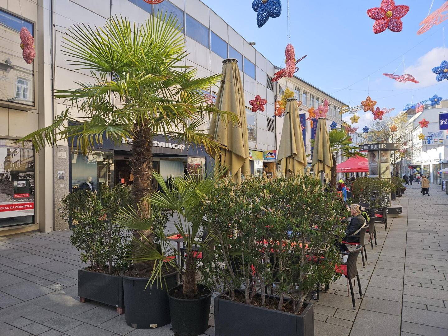 Belebtes Stadtbild einer Fußgängerzone mit Außengastronomie, Pflanzenkübeln und bunten Dekorationen.