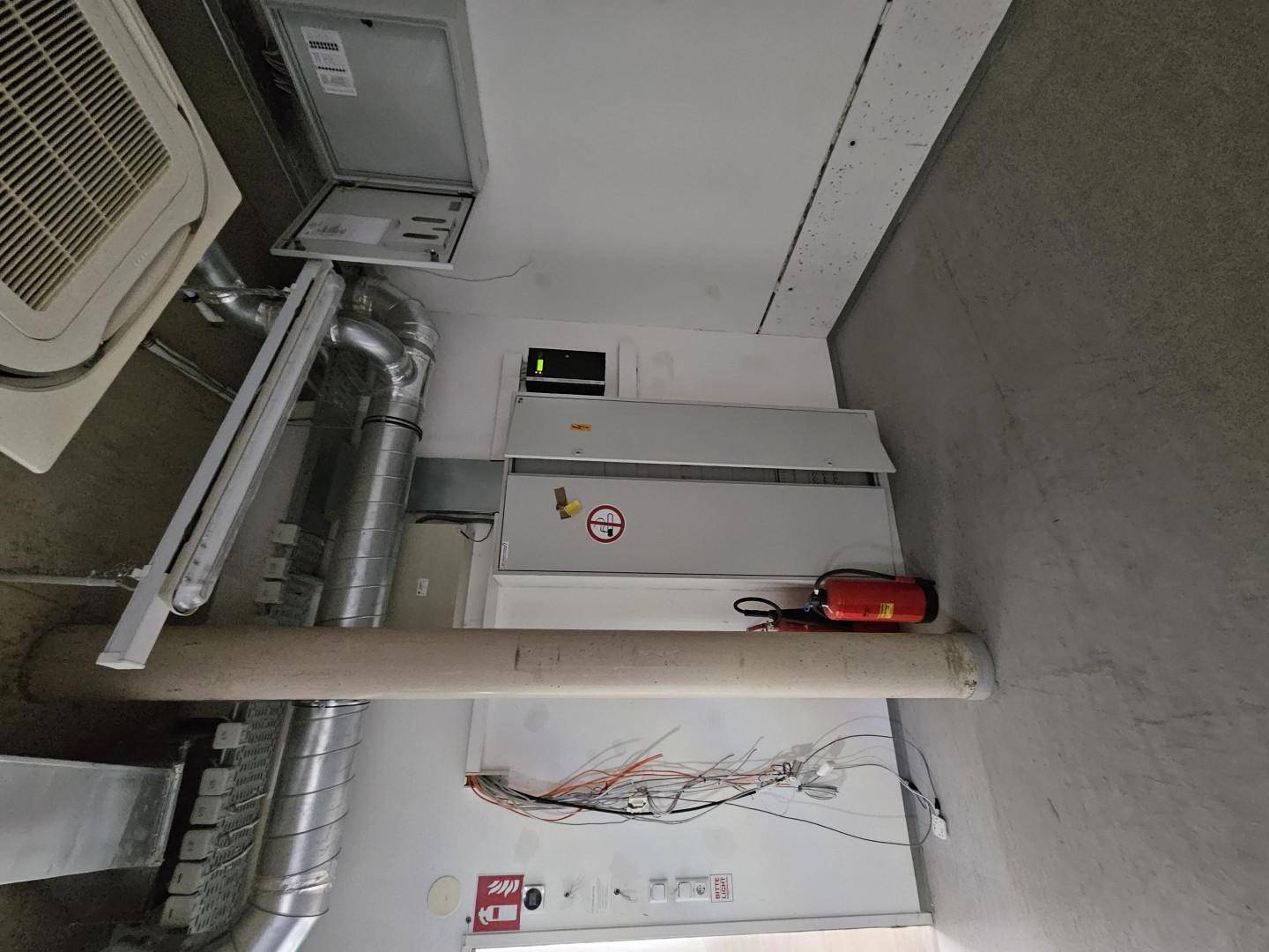 Technischer Raum mit sichtbaren Lüftungsrohren, elektrischen Installationen und einem Feuerlöscher an der Wand.