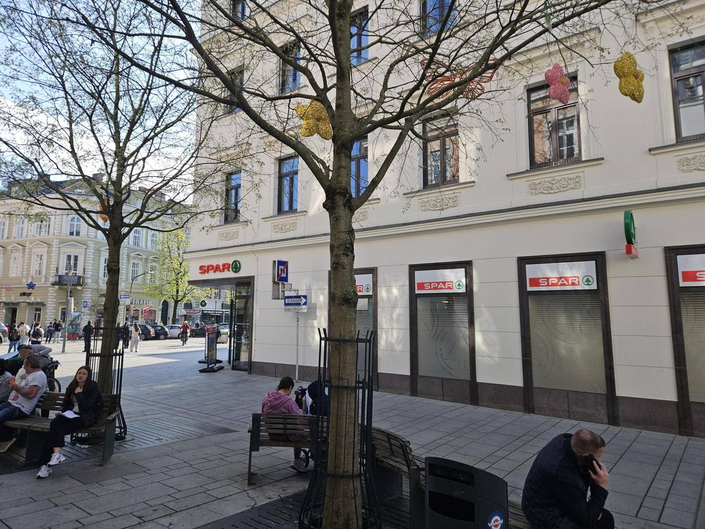 Ansicht einer belebten Fußgängerzone mit Geschäften, Sitzbänken und Bäumen unter blauem Himmel.