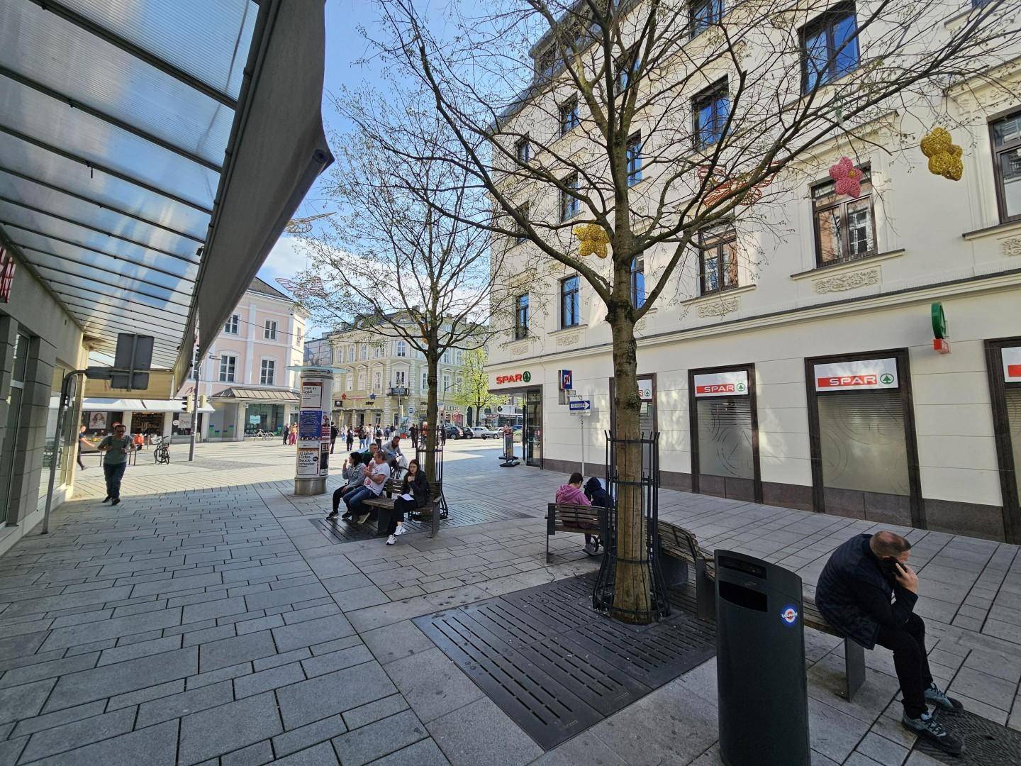 Weitläufige Ansicht einer Fußgängerzone mit Geschäften, Sitzgelegenheiten und Bäumen an einem sonnigen Tag.