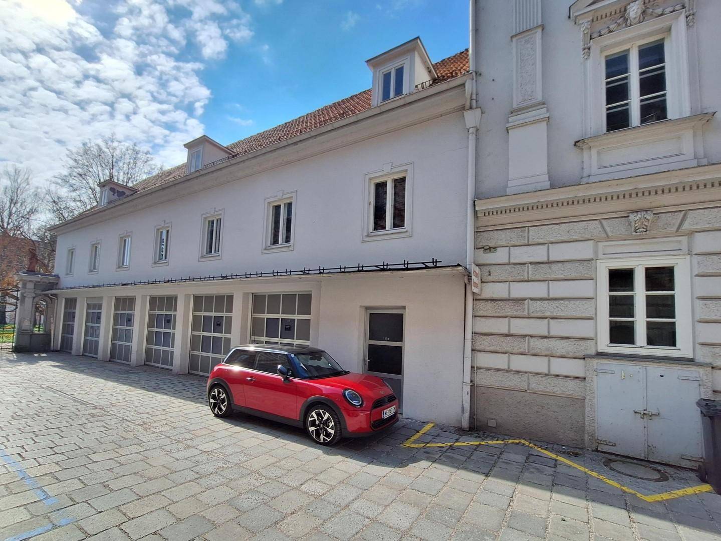 Seitenansicht eines Gebäudes mit mehreren Garagentoren und einem geparkten roten Kleinwagen auf dem Hof.