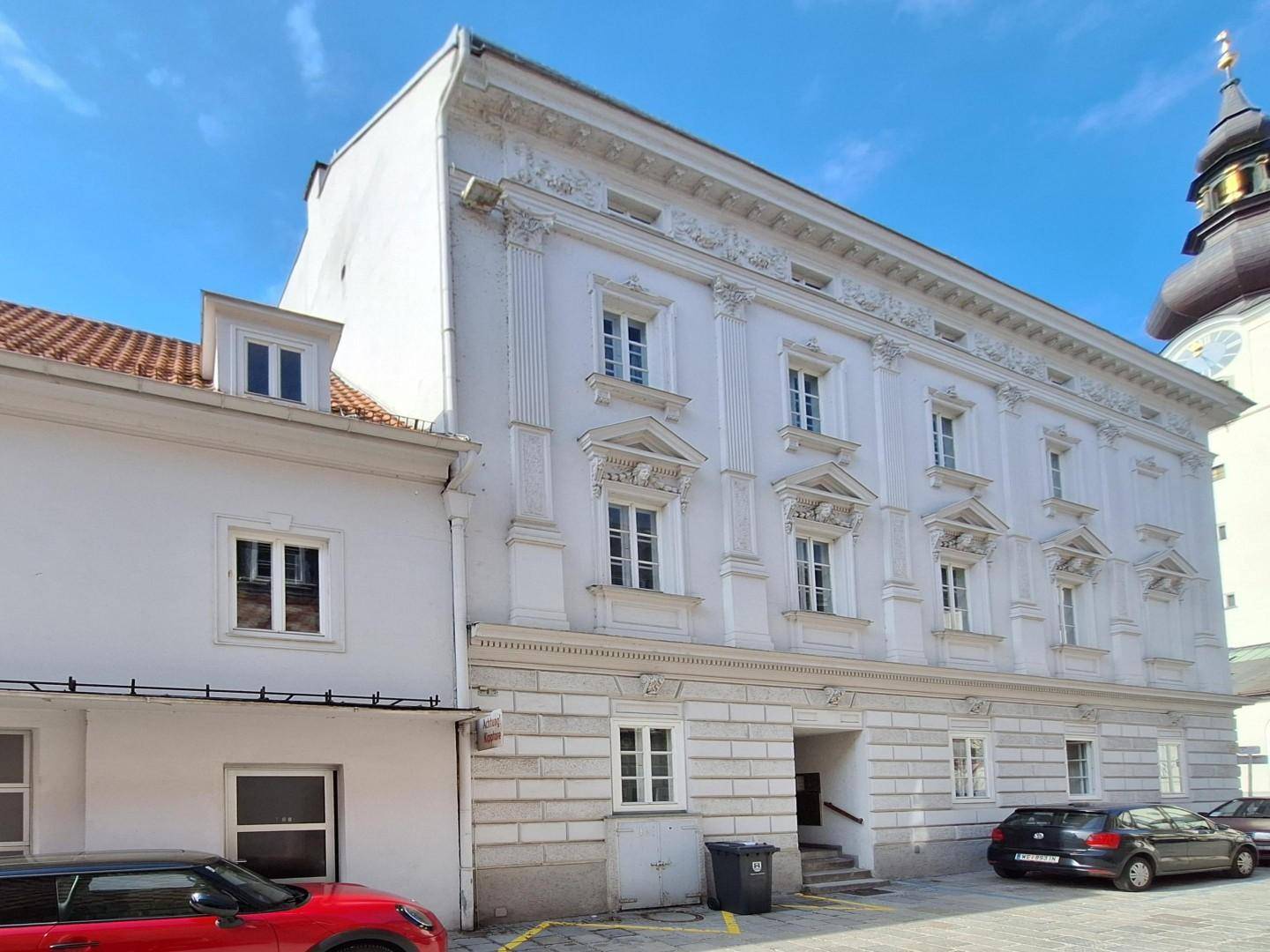 Historisches Gebäude mit weißer Fassade und einem angrenzenden Nebengebäude unter klarem Himmel.