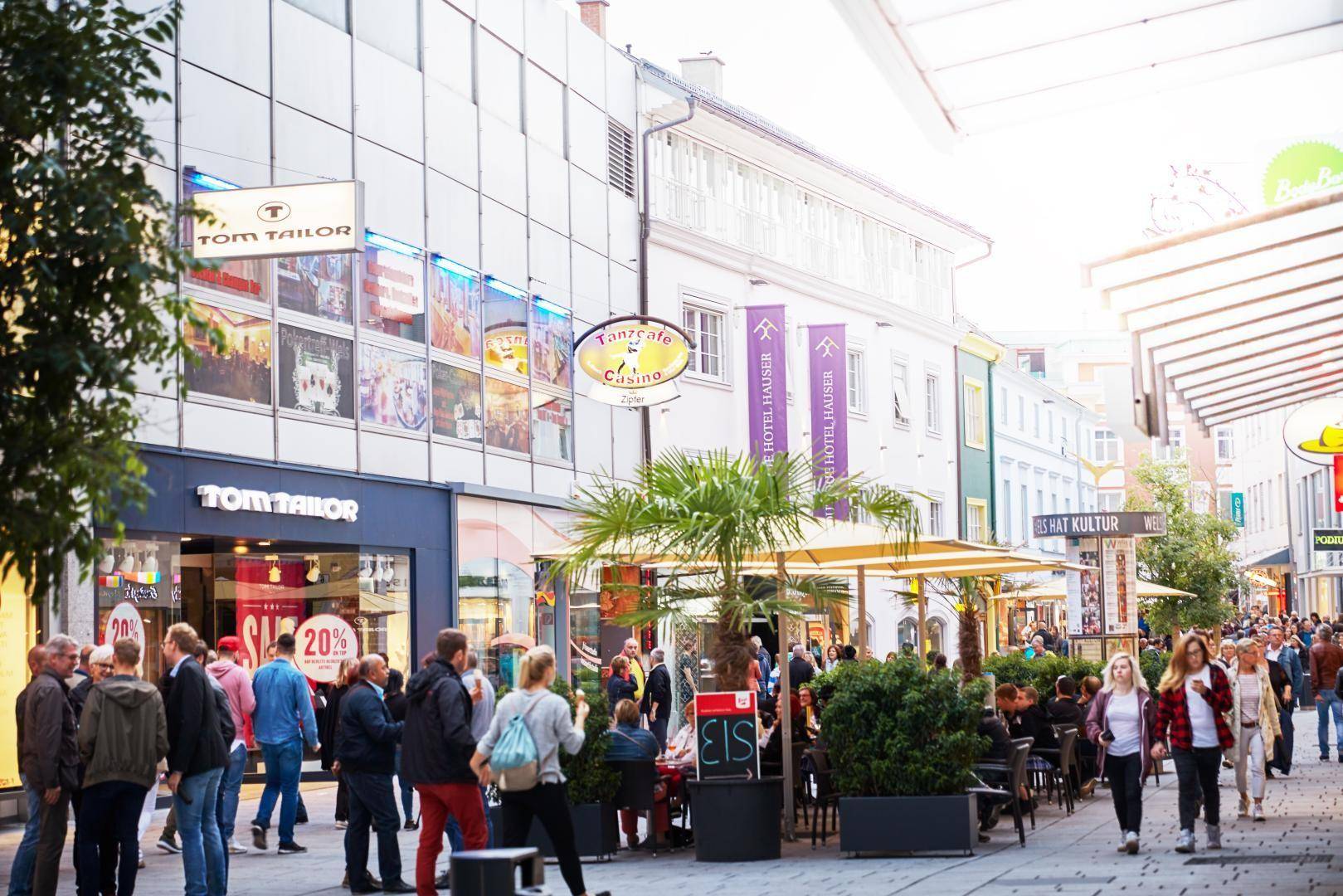Geschäftige Einkaufsstraße mit vielen Passanten, Geschäften und Außengastronomiebereichen.