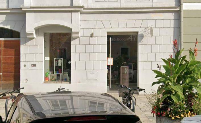 Straßenansicht eines Gebäudes mit großen Schaufenstern und einem modernen Eingang, teilweise verdeckt von einem Auto.