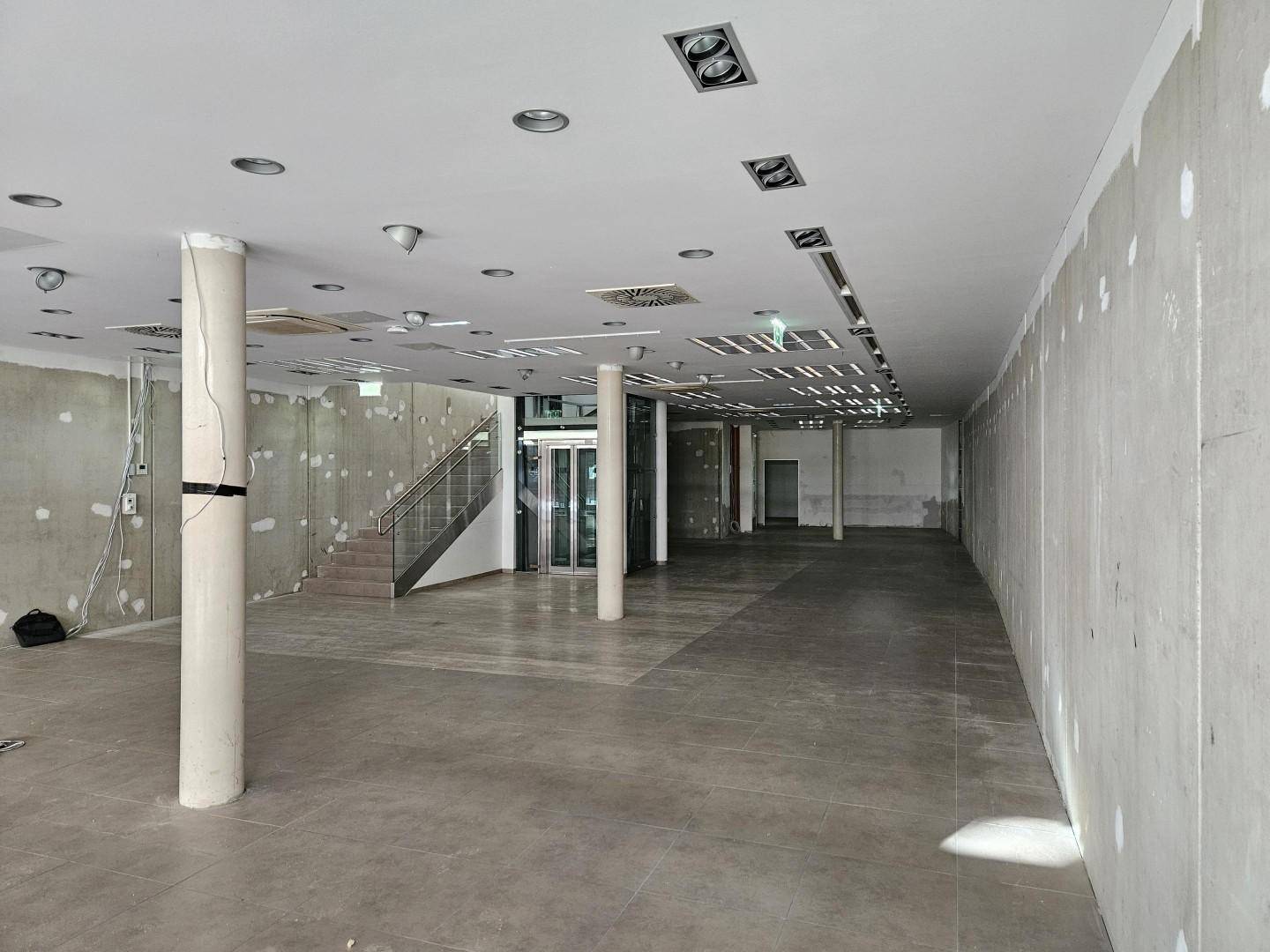 Weitläufiger Gewerberaum im Rohbau mit Säulen, Treppe und Glasaufzug, bereit für den Ausbau.