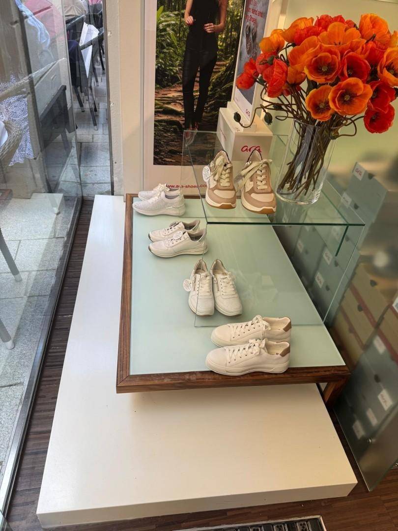 Nahaufnahme einer Schaufensterauslage mit weißen Sneakern und einem Blumenarrangement in einem Schuhgeschäft.