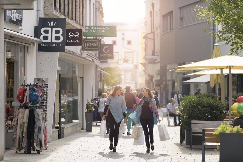 Belebte Einkaufsstraße mit Geschäften und Passanten, die bei Sonnenschein einkaufen gehen.