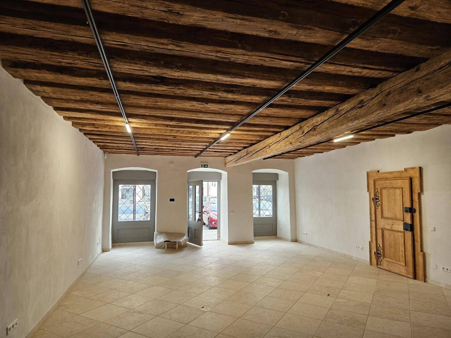 Geräumiger Innenraum mit sichtbaren Holzdeckenbalken, gefliestem Boden und mehreren Fenstertüren zur Straße.
