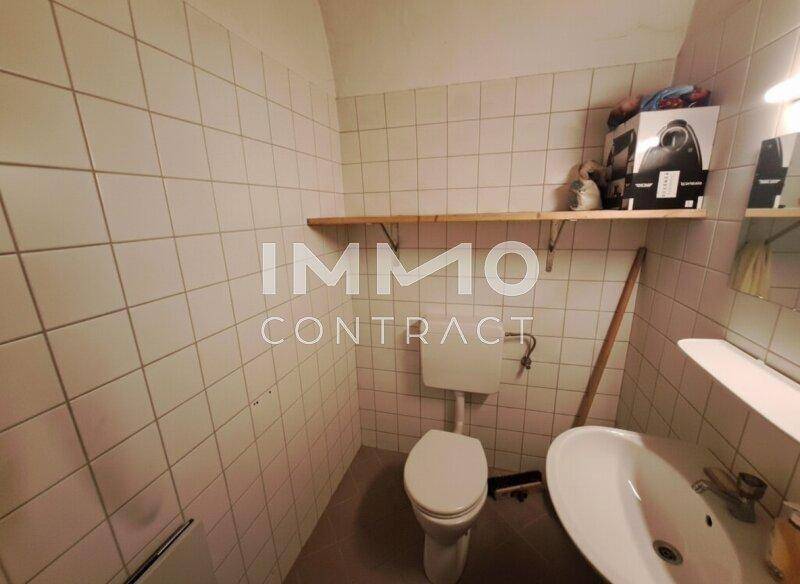 Kleiner Toilettenraum mit weißen Fliesen, einem Waschbecken und einer einfachen Holzablage.
