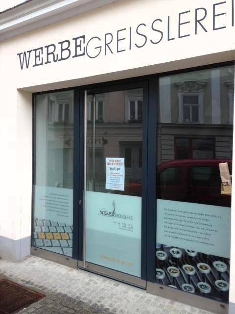 Eingangsbereich eines Geschäfts mit moderner Glasfront und dem Schriftzug 'WERBEGREISSLEREI' an der Fassade.