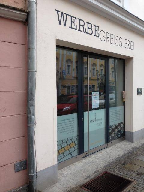 Außenansicht eines Geschäftsgebäudes mit moderner Glasfront, Regenfallrohr und dem Schriftzug 'WERBEGREISSLEREI'.