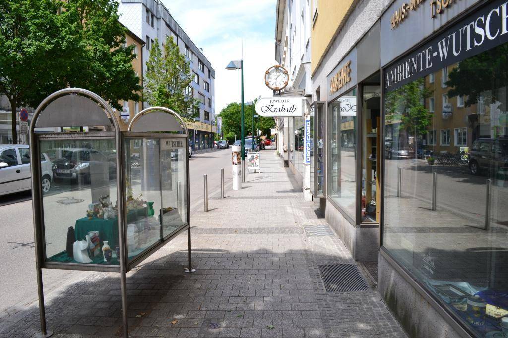 Belebte Geschäftsstraße mit verschiedenen Ladenlokalen, parkenden Autos und Bäumen an einem sonnigen Tag.