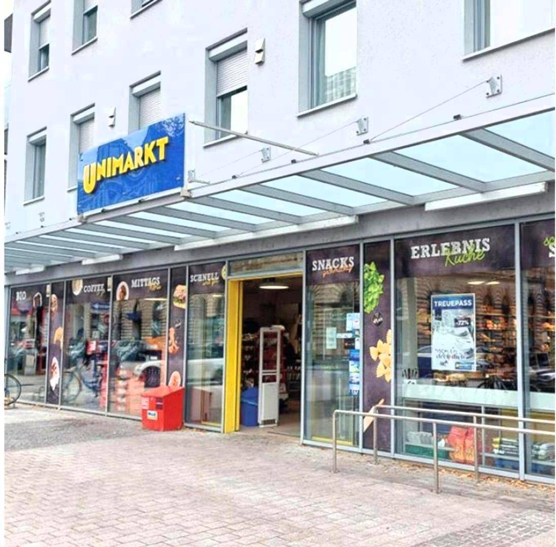 Eingangsbereich eines Supermarktes mit großen Schaufenstern, einem Vordach und dem "UNIMARKT"-Schild.
