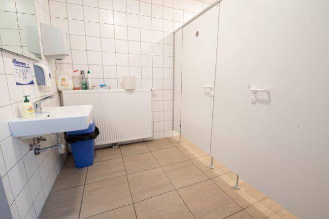 Einfacher Toilettenraum mit weißen Fliesen, Waschbecken und mehreren Kabinen.