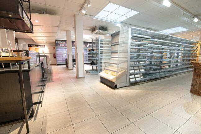 Blick in einen Supermarktgang mit leeren Regalen, einer Frischetheke und Deckenbeleuchtung.