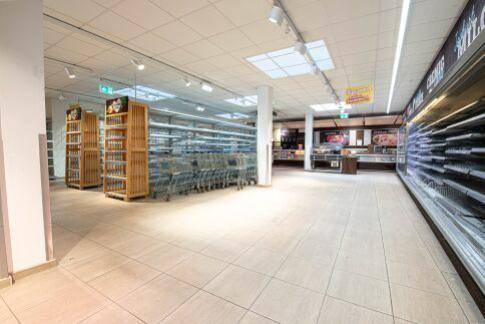 Weiterer Blick in einen leeren Supermarktgang mit Regalen, Einkaufswagen und Deckenbeleuchtung.