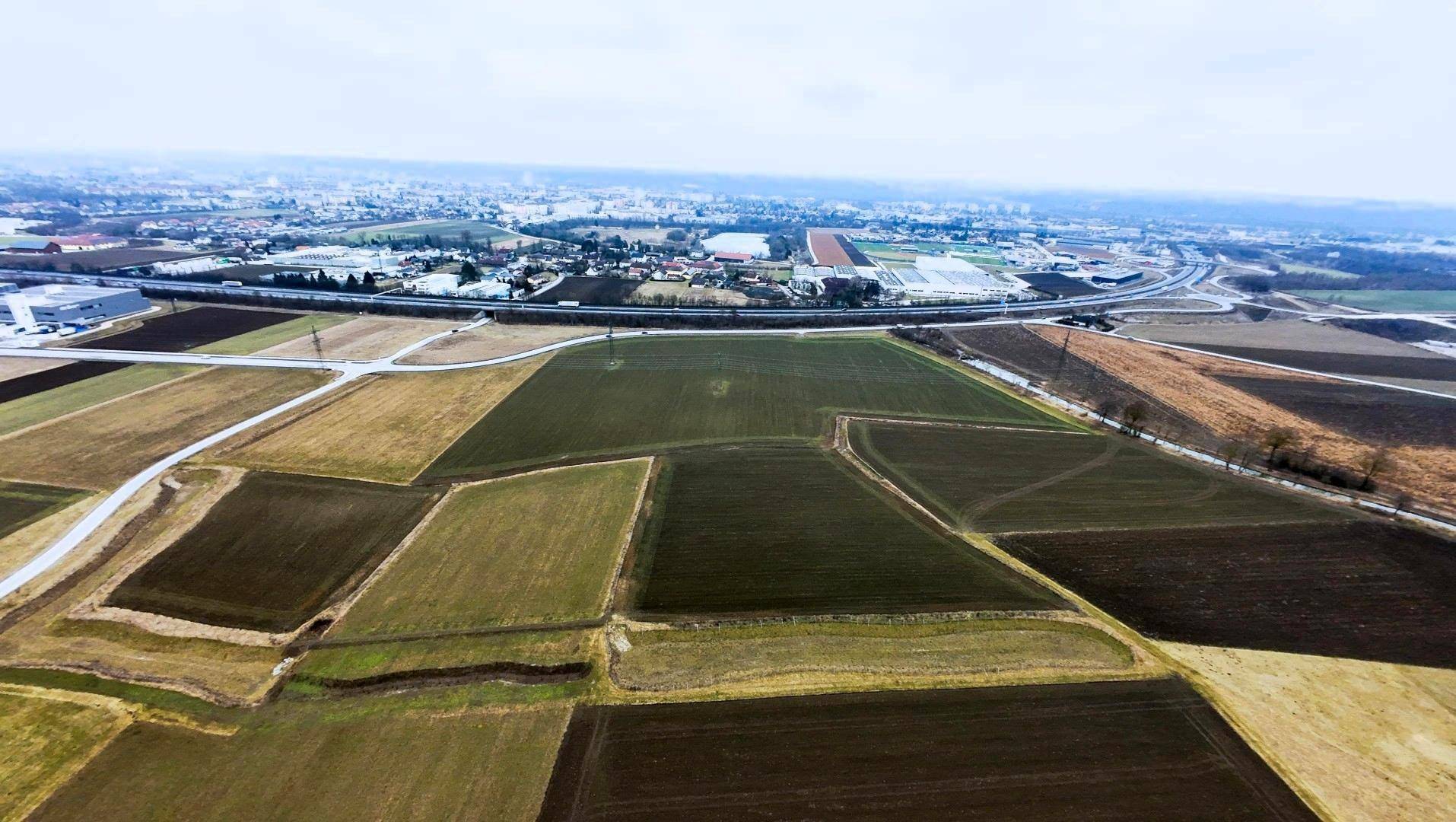 Luftaufnahme einer weiten Landschaft mit Feldern, Straßen und einer Stadt in der Ferne unter bewölktem Himmel.