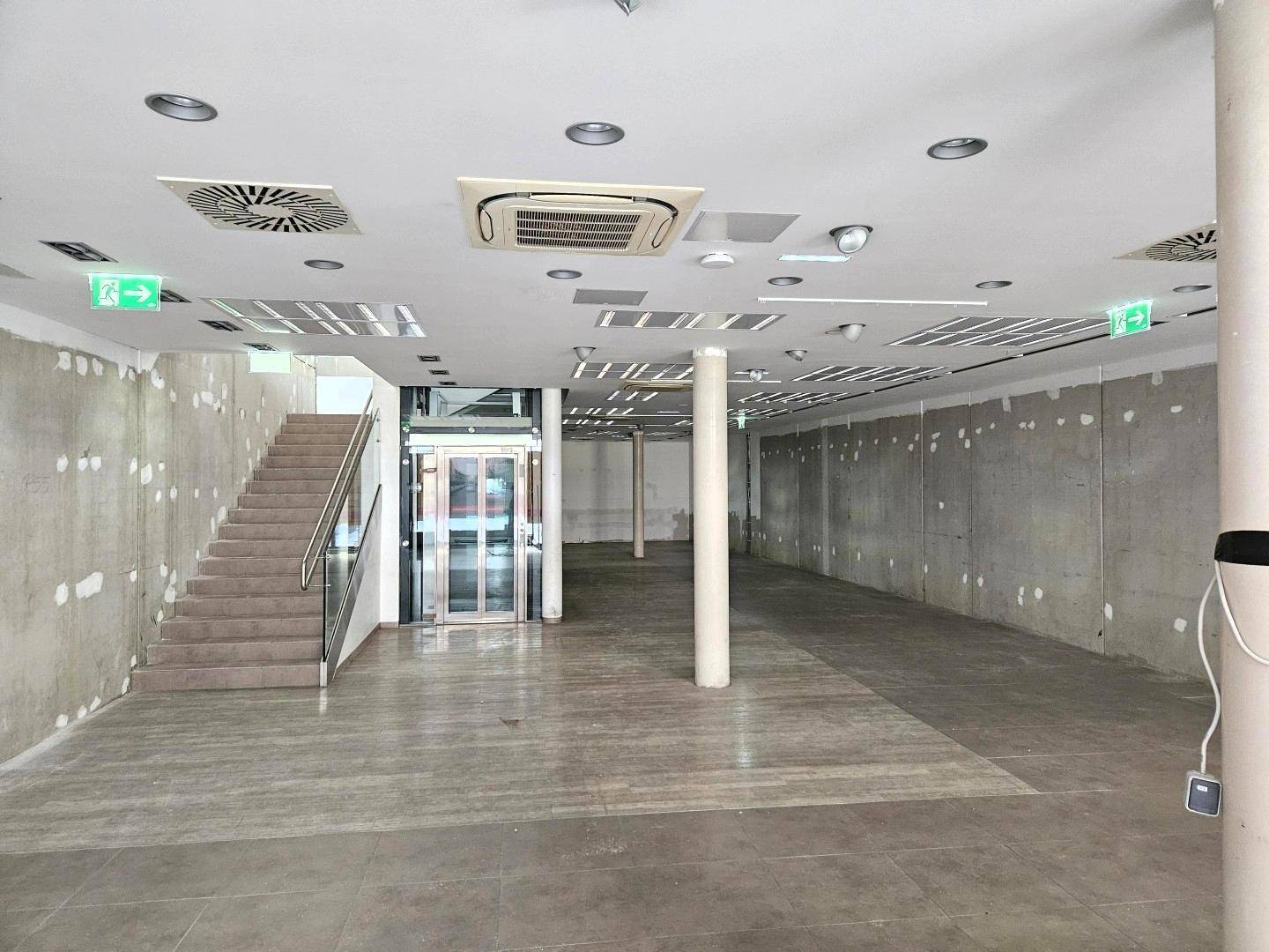 Geräumiger, unfertiger Gewerberaum mit Betonboden, Treppe, Glasaufzug und sichtbaren Lüftungsanlagen an der Decke.