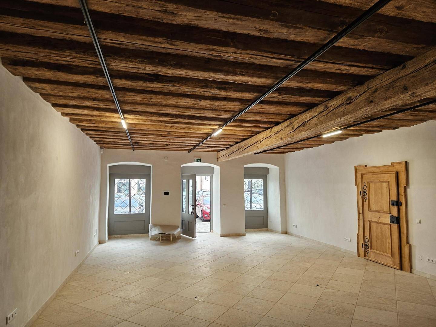 Heller Wohnraum mit sichtbaren Holzbalken und großen Fenstern, die viel Tageslicht hereinlassen, auf Fliesenboden.